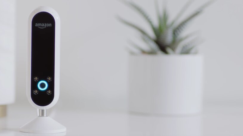 The Echo Look, a pill-shaped black and white camera with four flash sensors around a central camera surrounded by a blue illuminated Alexa light ring. Shown on a table.