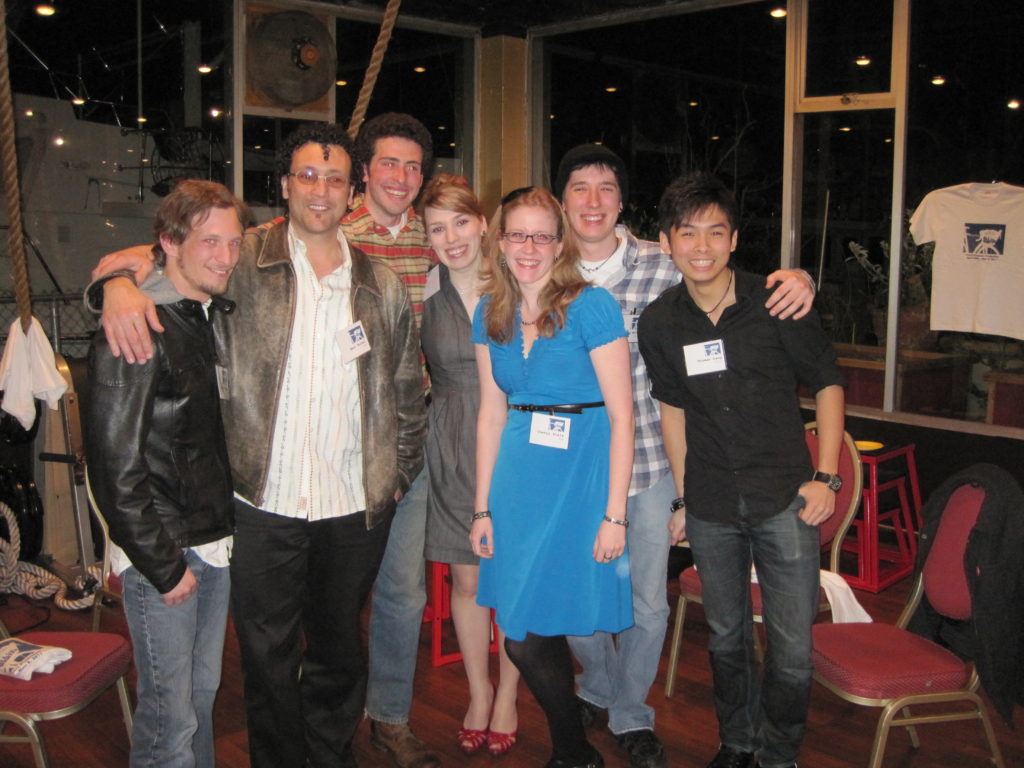 A group of smiling people of various ages and races hugs and looks directly at the camera, surrounded by T-shirts with the "All I Ever Wanted" logo of a bike with a basket.