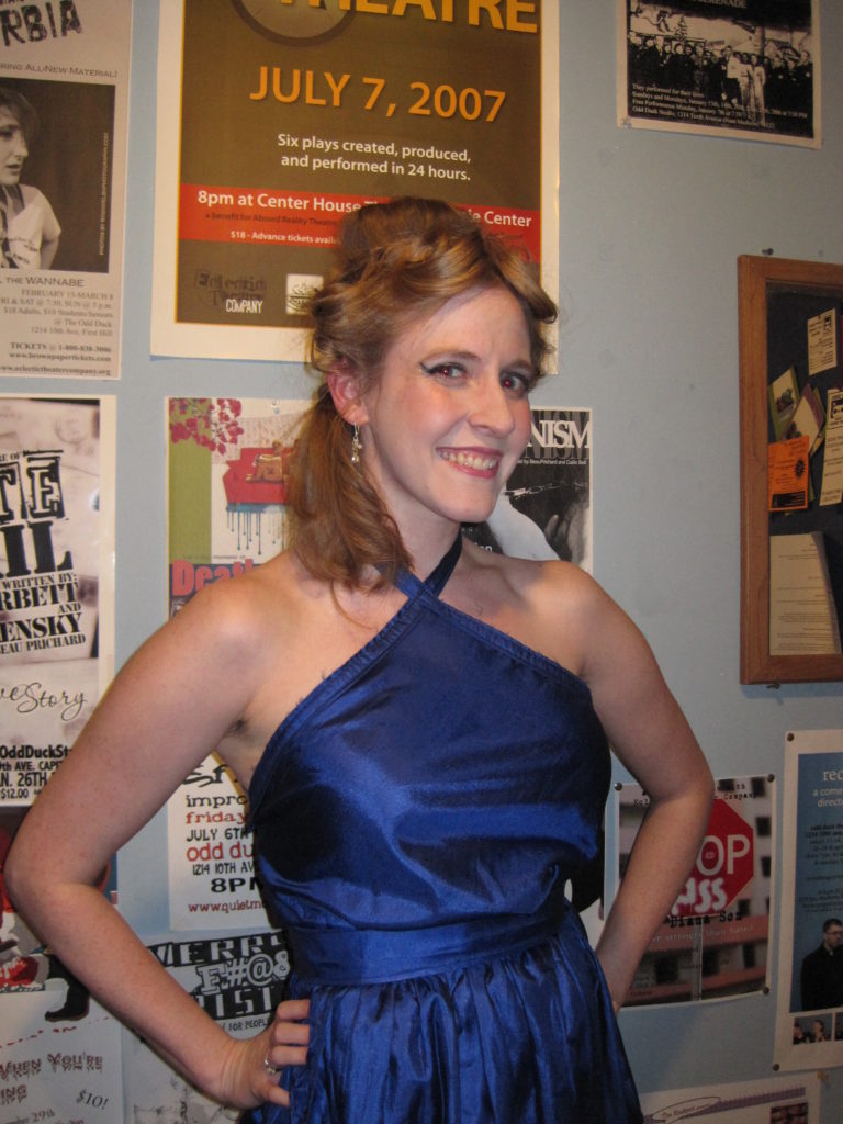 Cheryl poses in front of a wall of show posters with a space age hairdo and a blue halter dress.