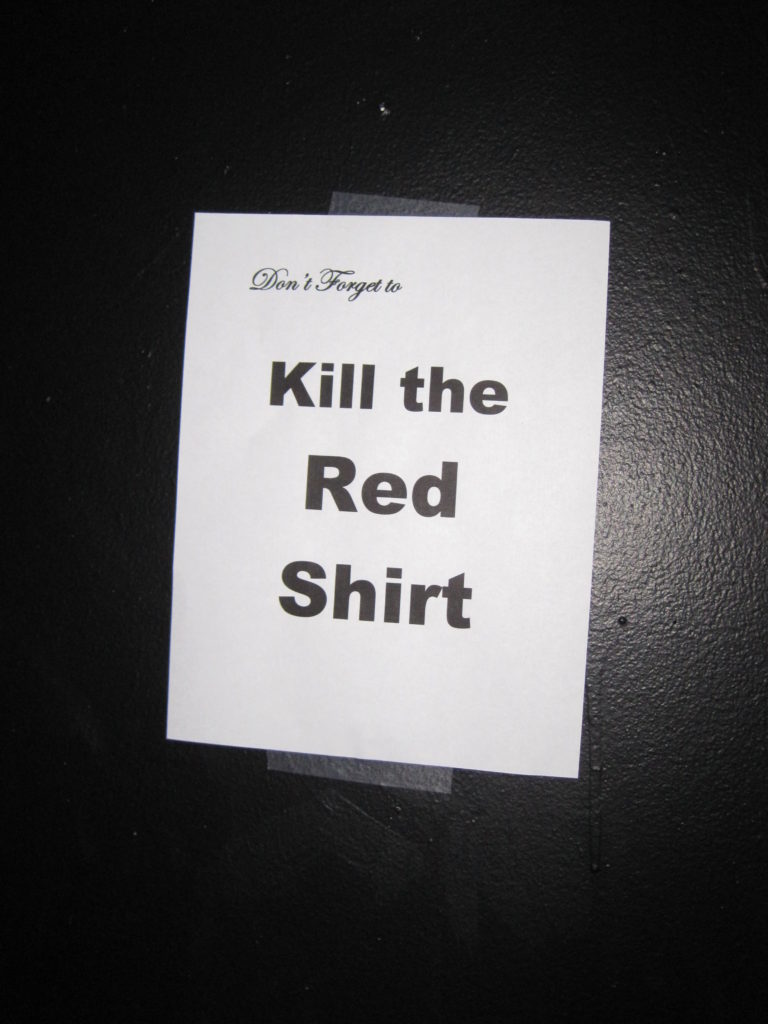 A white sheet of paper on a black backstage wall that reads in laser printed lettering "Don't forget to... Kill the Red Shirt"